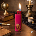A pink refillable lighter with a flame lit, standing on a wooden desk. The lighter features an illustration of a hand holding a cup and has a black top with a gold button.
