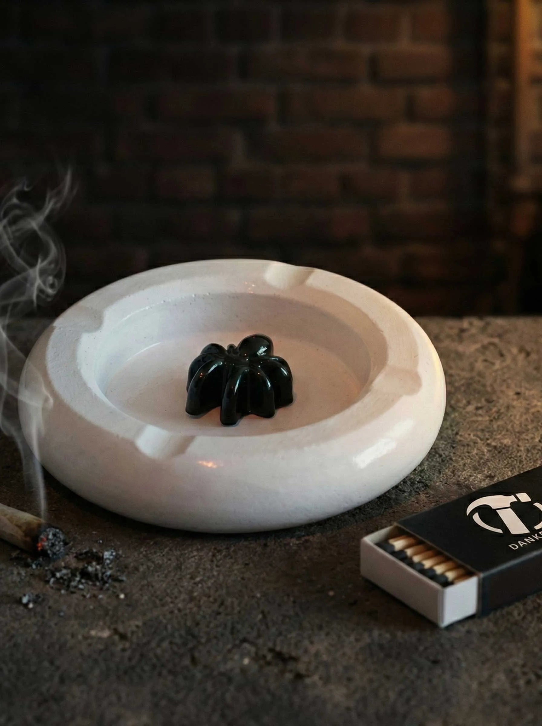 White concrete ashtray with a black spider decoration on a dark surface, with a matchbox and matches nearby.