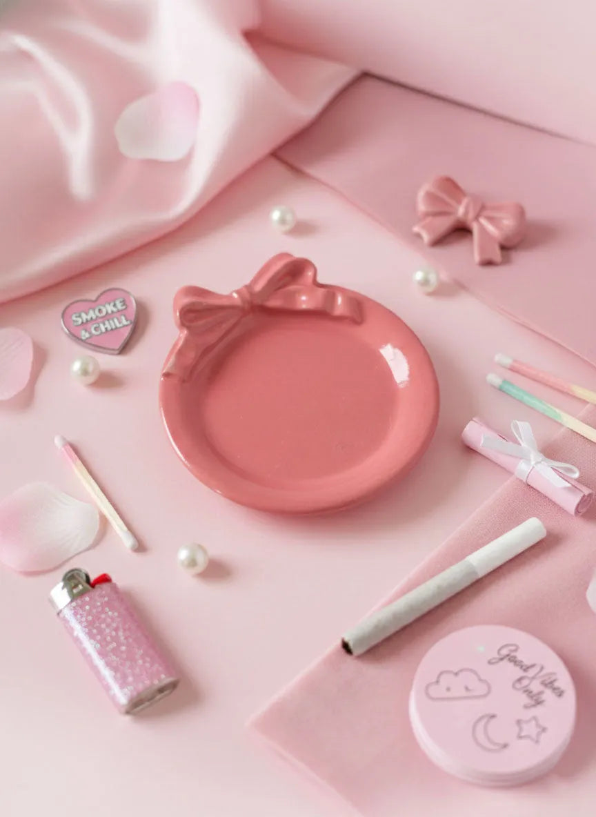 Pink Pooki puff ashtray including a plate, lighters, and small objects on a pink bedsheet background.