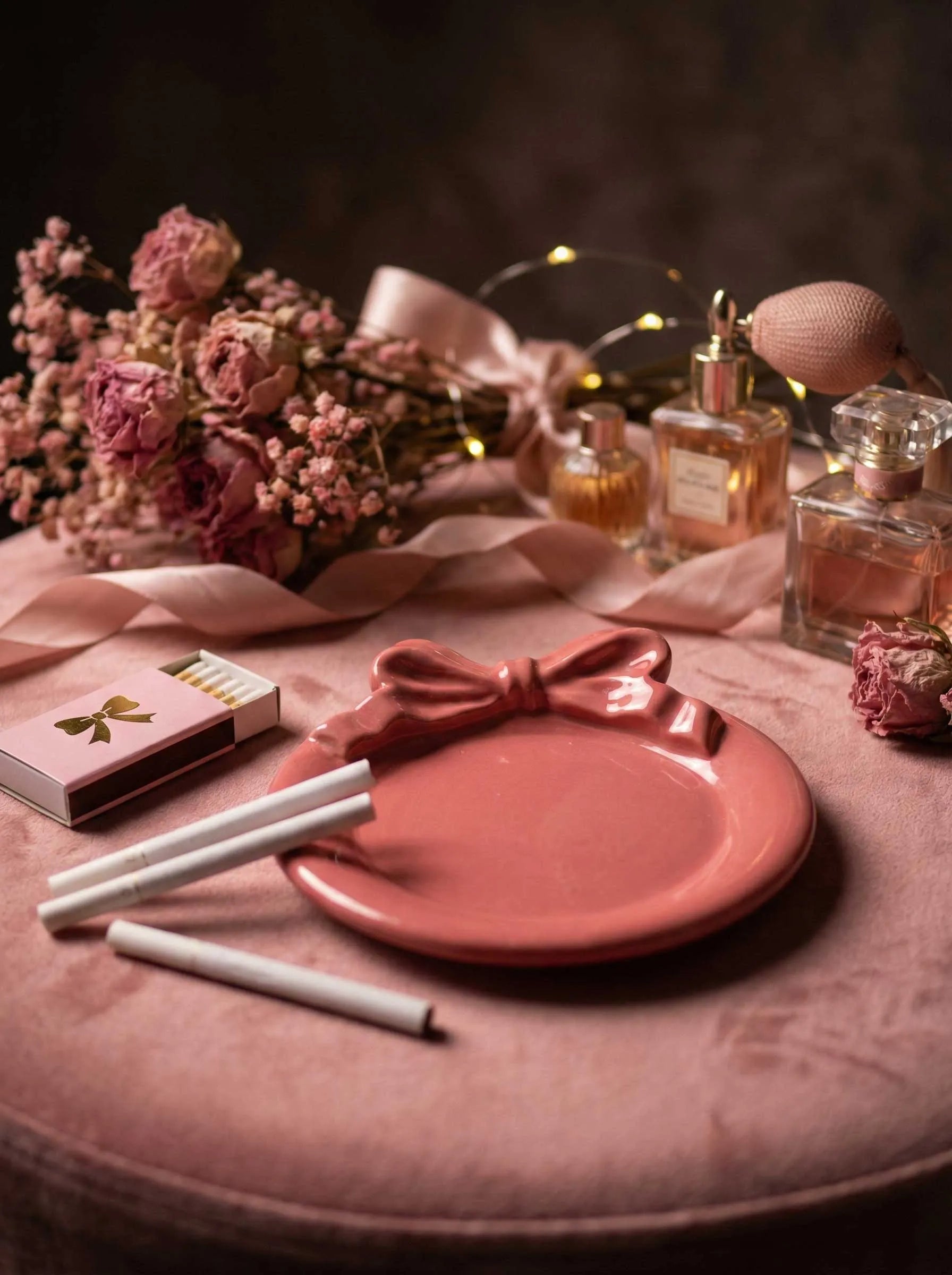 Pink plate ashtray with a bow design, cigarettes, and perfume bottles on a table with flowers and lights.