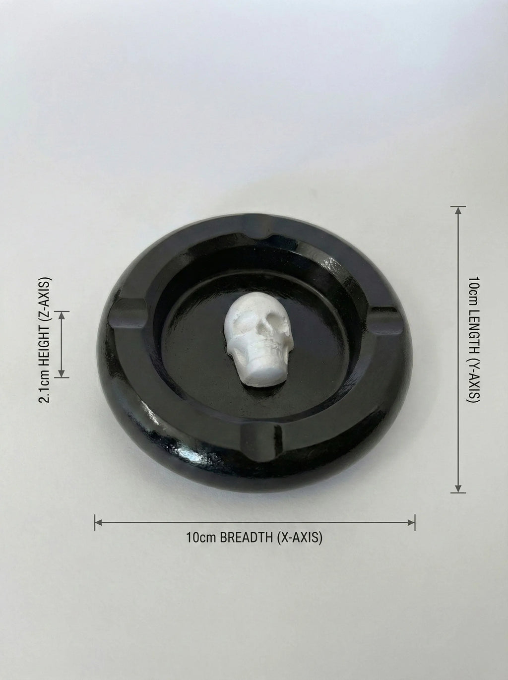 Black circular ashtray with a white skull-like figure inside, on a light gray background with measurements indicated.