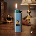 A light blue and beige lighter with a cartoon angel graphic and the text 'STONED PARI' stands lit on a wooden table, surrounded by a lamp, books, and a cup in a warm, vintage-style setting.