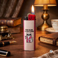 A pink and white cigarette lighter with a funky cat wearing sunglasses and the text 'TOO GLAM TO GIVE A DAMN' stands lit on a wooden desk surrounded by vintage books, a lamp, and stationery.