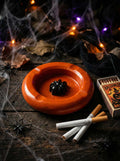 Halloween-themed ashtray with spider design on a dark surface with matches and cigarettes.